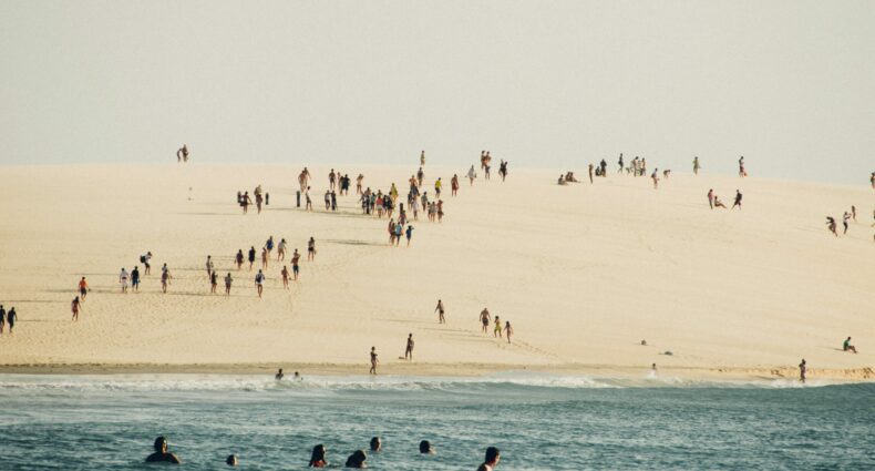 A foto mostra uma praia em Jericoacoara, com pessoas descendo e subindo as dunas de areia. Mostra também uma porção do mar, onde há pessoas nadando.