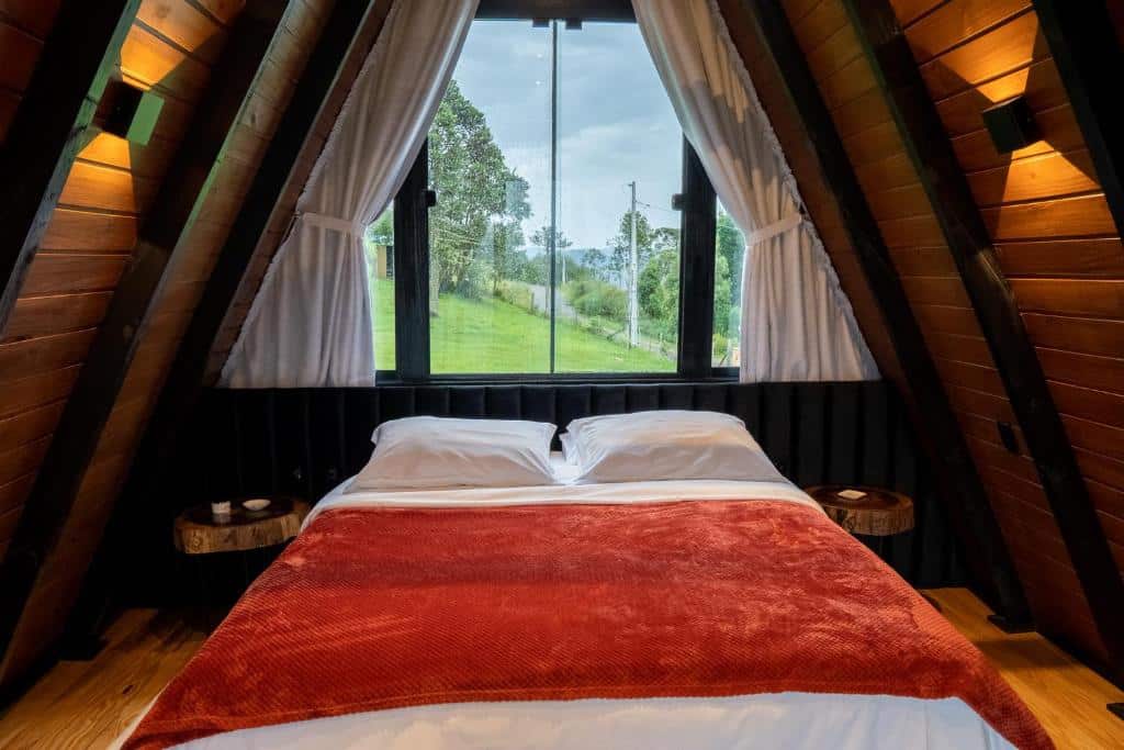 Quarto do Altitude 1140 Urubici - Chalés com cama de casal ao centro, mesinhas de madeira de ambos os lados e uma janela de vidro e madeira bem em cima da cama, com vista da natureza.
