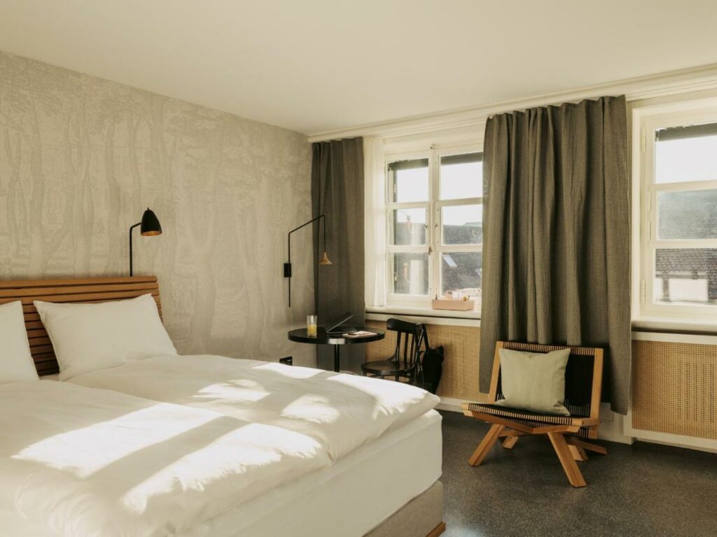 Imagem do quarto do Boutique & Design Hotel Volkshaus Basel com cama de casal do lado esquerdo da imagem do lado esquerdo da cama uma mesa redonda com uma cadeira e do lado direto dessa mesa uma outro poltrona com uma almofada em cima.