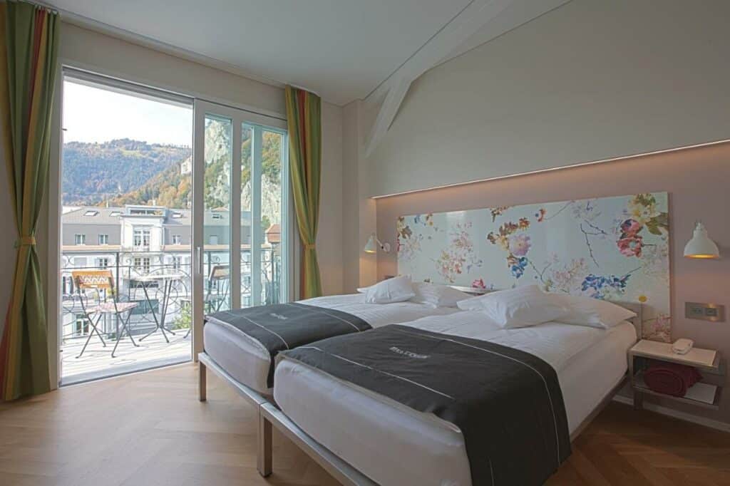Quarto do Boutique Hotel Bellevue com cama de casal do lado direito da imagem no centro do quarto e em cada lado da cama uma cômoda e do lado direito da cama uma porta que dá acesso a varanda. Representa hotéis em Interlaken.