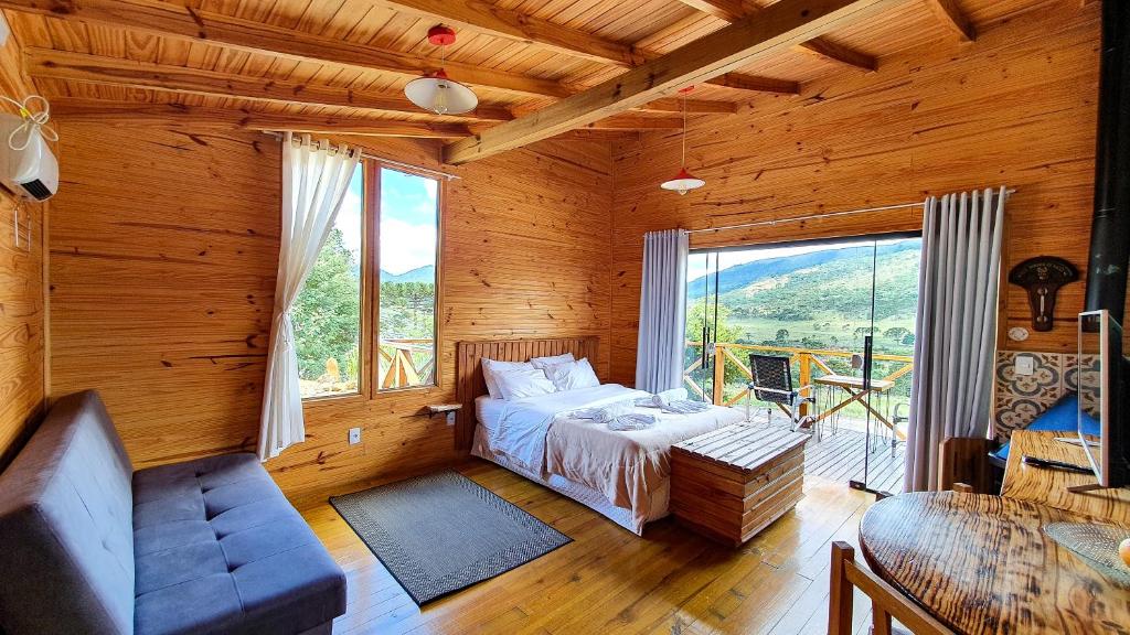 Quarto de um dos chalés em Urubici, a Cascata dos Amores, com cama de casal com um baú de madeira à frente, há ainda mesa redonda com cadeiras também em madeira, tv e um sofá de um lado do quarto, do outro, há uma varanda com cadeiras e vista da natureza.