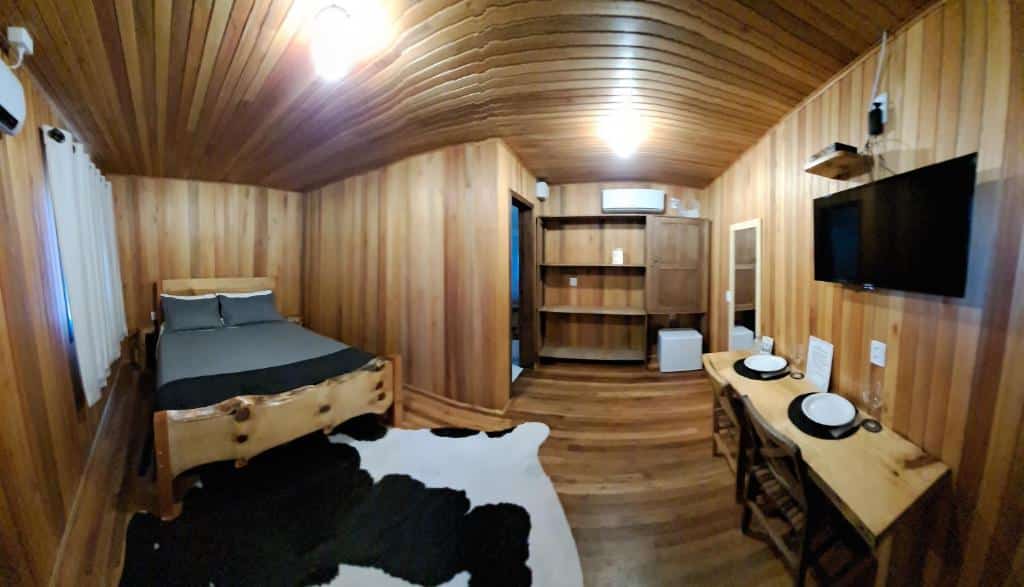 Quarto do Chalés Sossego da Serra em Urubici, com cama de casal de um lado, mesa com cadeiras e tv em frente, tapete que imita pele de vaca no chão, e há ainda prateleiras do outro lado.