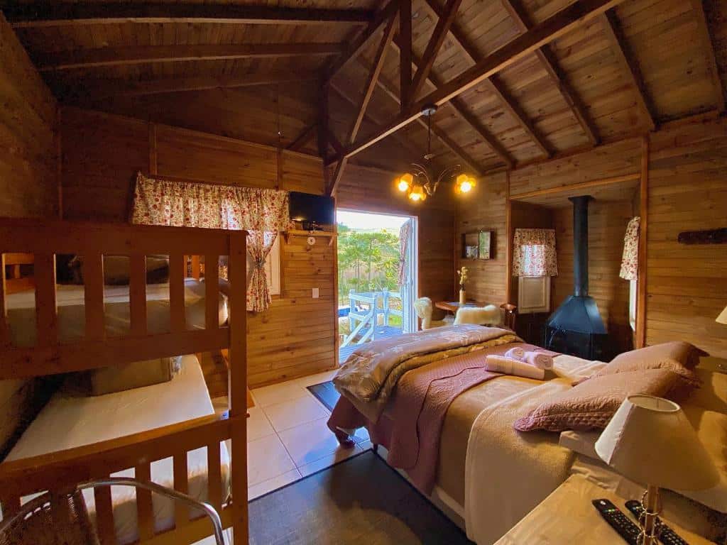 Quarto do Estalagem Villa Serrana, com cama de casal ao centro e uma beliche mais perto da parede, há luzes no teto, janela e porta grande com vista da natureza, a decoração é rústica.