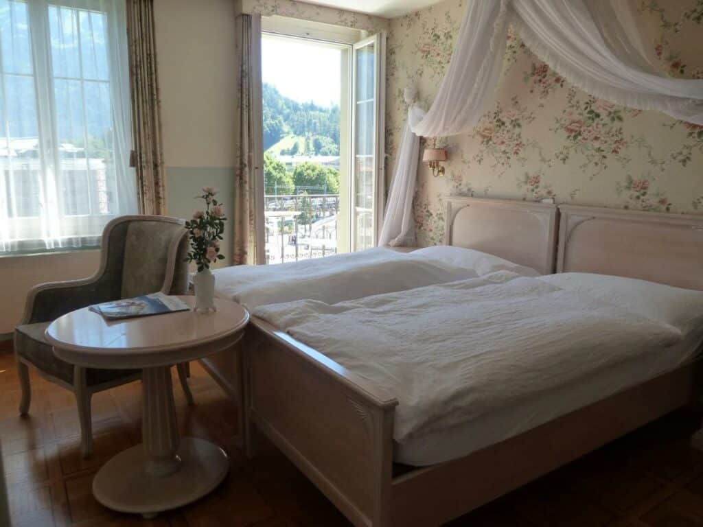 Quarto do Hotel Central Continental com cama de casal do lado direito da imagem no centro do quarto nos pés da cama uma mesa redonda com uma cadeira e do lado direito da cama um porta que dá acesso a varanda.