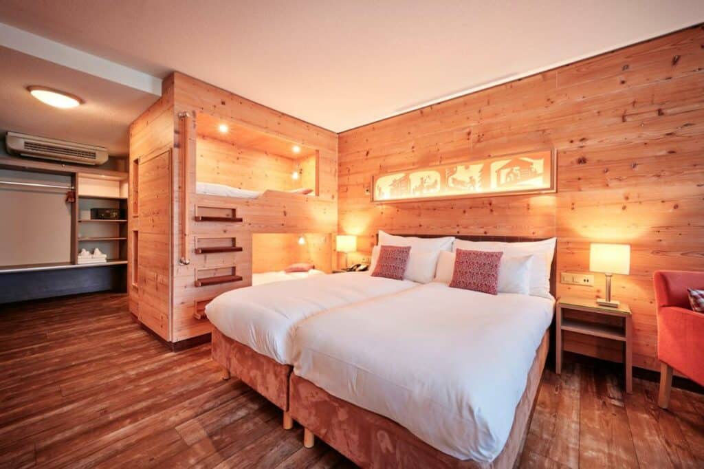 Imagem do quarto do Hotel Interlaken com cama de casal do lado no centro do ambiente em cada lado do quarto uma cômoda com luminária e do lado direito da cama ao lado da cômoda uma beliche. Representa hotéis em Interlaken.