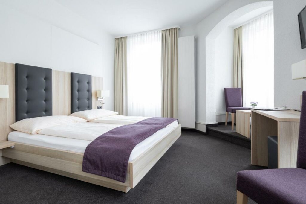Quarto do Hotel Lötschberg com cama de casal do lado esquerdo da imagem em frente a cama uma mesa de trabalho e uma TV presa na parede do lado esquerdo do ambiente uma poltrona com uma mesinha. Representa hotéis em Interlaken.
