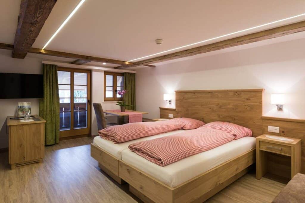 Quarto do Hotel Restaurant Hirschen com cama de casal do lado direito da imagem no centro do quarto em cada lado da cama uma cômoda do lado direito da cama uma mesa com cadeira e ao fundo uma cômoda e uma TV presa na parede. Representa hotéis em Interlaken.