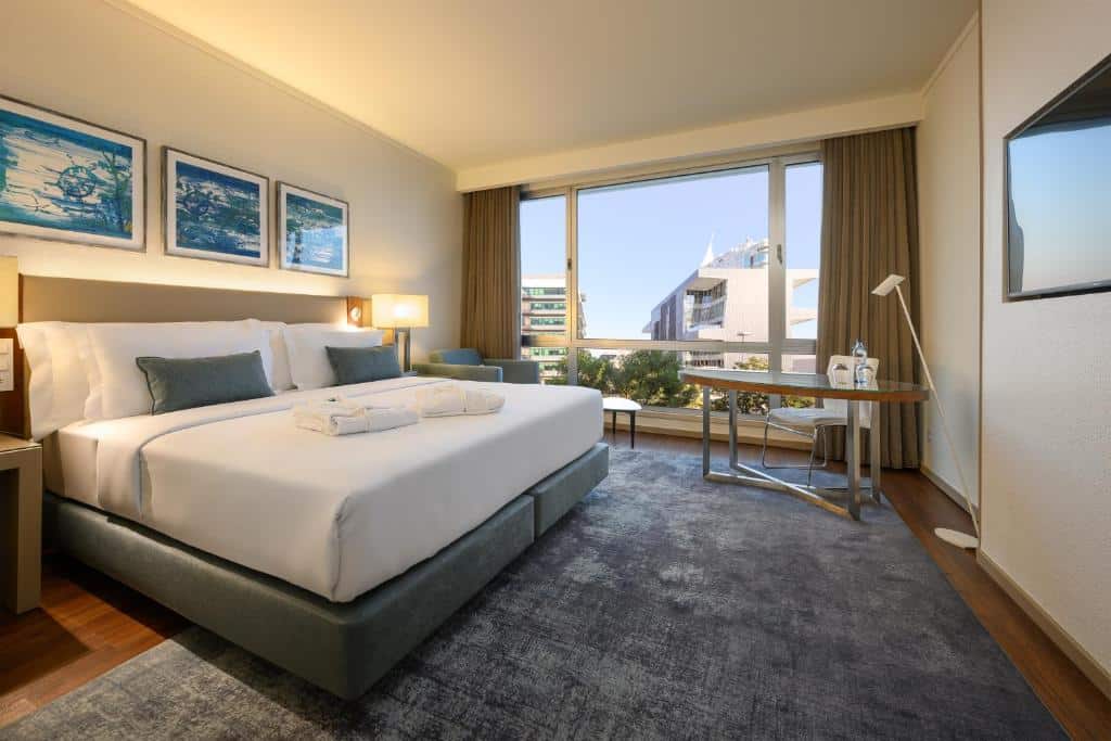 Quarto do Melia Lisboa Oriente Hotel com uma cama de casal com travesseiros e almofadas, há uma janela ampla com cortinas, uma mesa com uma cadeira e uma luminária de chão, com uma televisão de frente para a cama, há também alguns quadros na parede e um amplo tapete.