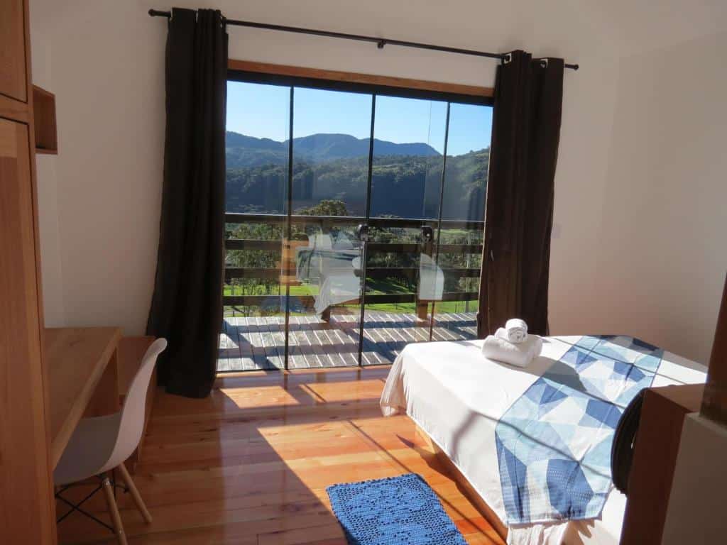 Quarto do Refúgio da Montanha, um dos chalés em Urubici, com cama bem em frente a uma porta de vidro que dá para uma varanda com mesinhas e cadeiras com vista da natureza, há cortinas pesadas e uma poltrona dentro do quarto.