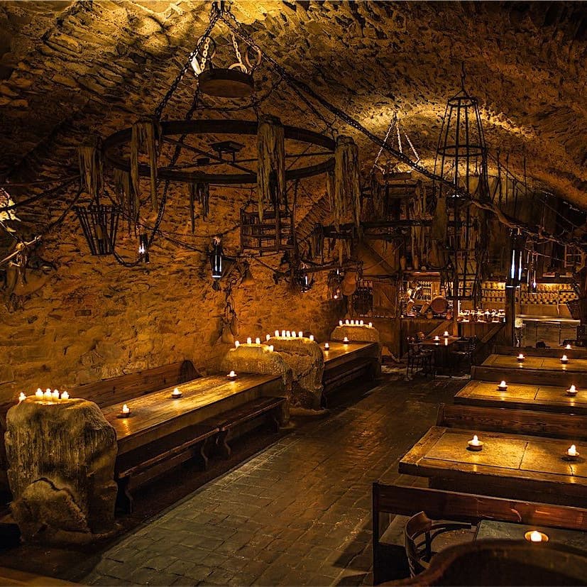 Imagem do interior do Krčma U Pavouka com mesas de madeiras no ambiente e velas em cima das mesas e bancos de madeiras.