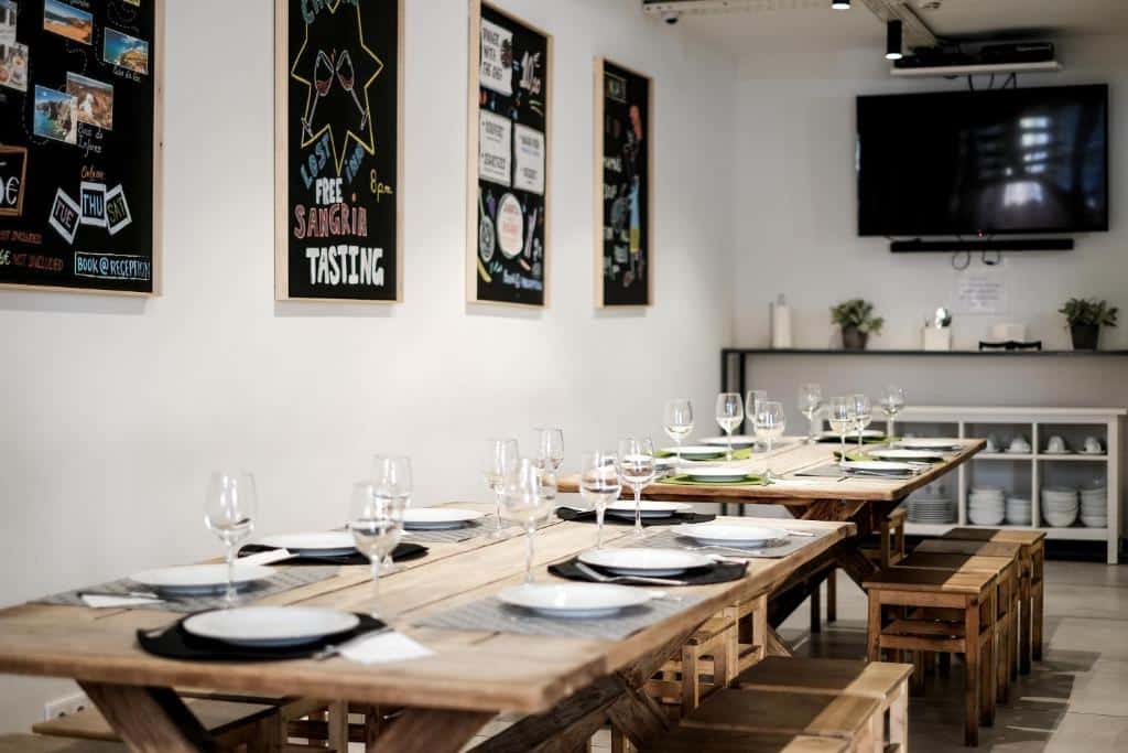 Sala de refeições do Lost Inn Lisbon Hostel com duas mesas grandes de madeira junto de bancos de madeira também, há uma televisão no local e alguns quadros na parede