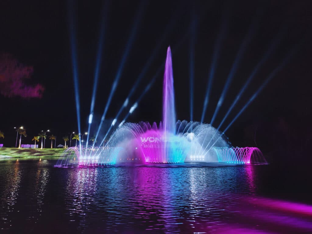 Um show noturno de fontes de água com jatos iluminados em roxo, rosa e azul, cercados por feixes de luz branca.