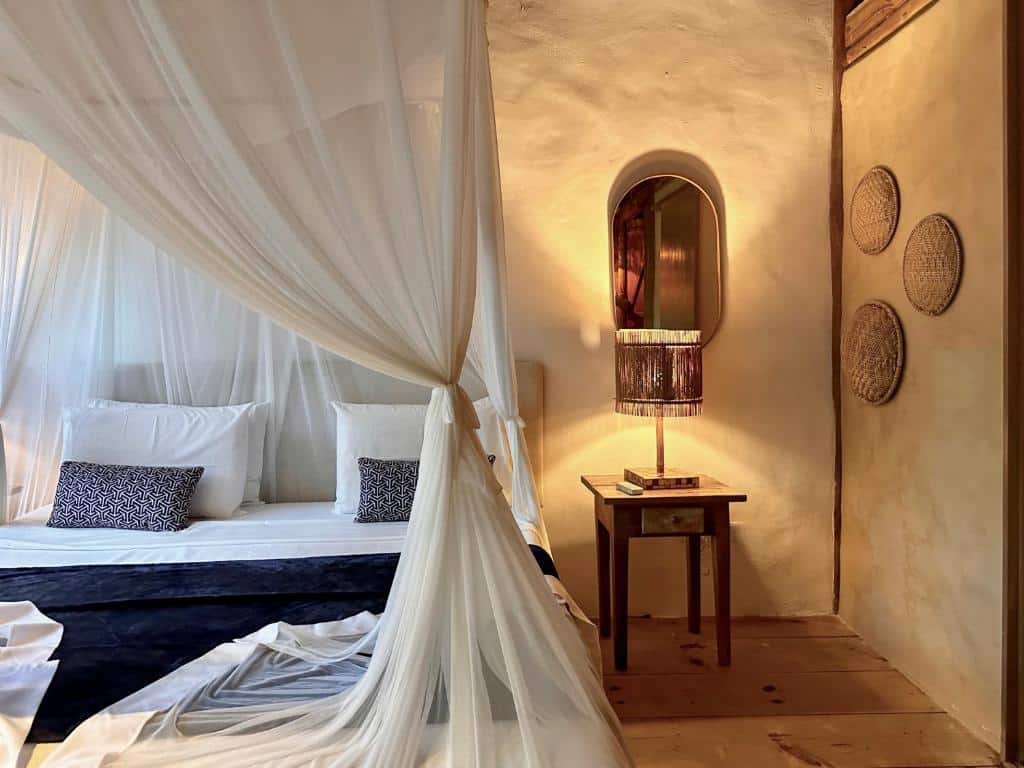 Foto que foca na cama da Soleluna Casa Pousada, em Trancoso. A cama de casal ocupa a esquerda da fotografia. Possui lençóis brancos e uma manta na cor azul marinho. Há tecidos sobre a cama formando uma espécie de tenda. Na direita vemos uma mesa de cabeceira rústica com abajur em cima, e um espelho. Também há algumas decorações de palha na parede direita.