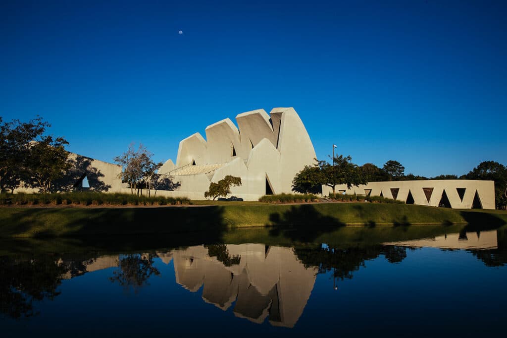 Foto tirada do Teatro L'Occitane, em Trancoso. Em primeiro plano há um lago, e o teatro está logo atrás, sobre um gramado verde. Sua arquitetura é toda branca e possui estilo contemporâneo. Há algumas árvores em volta dele. O céu está na cor azul escura, quase anoitecendo.