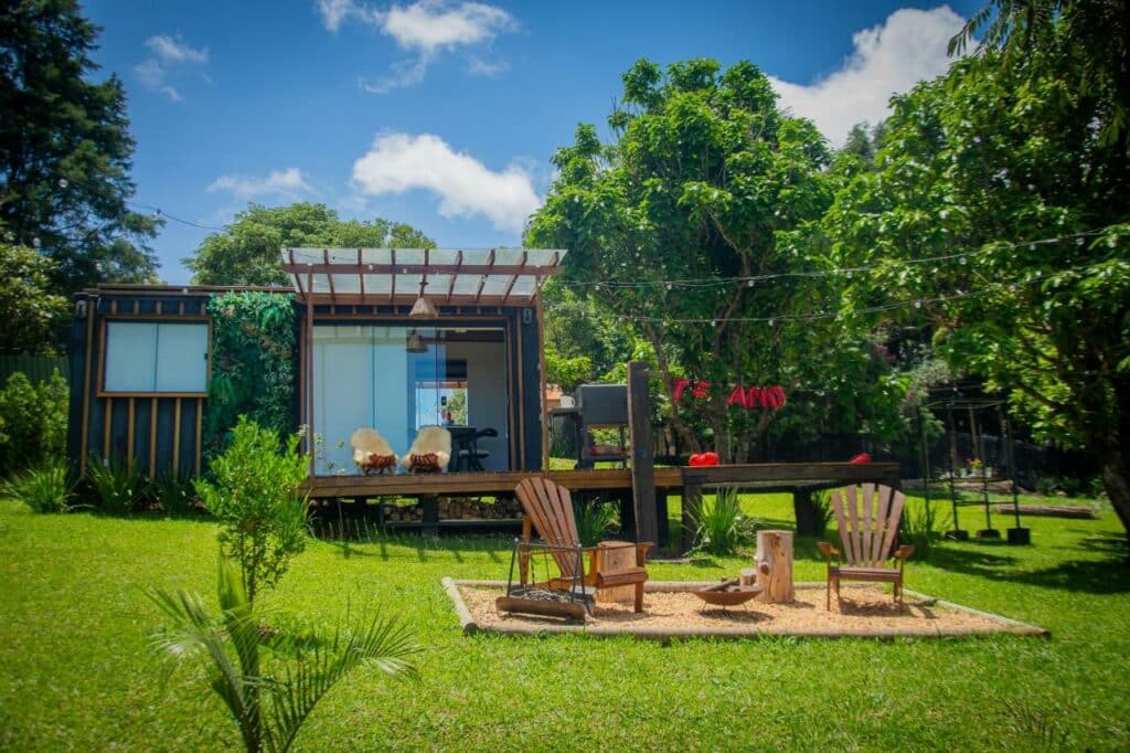 Frente do T24 Tinyhouse Container para ilustrar post sobre chalés em Ibiúna. Do lado direito uma área com areia e cadeiras, no fundo o deck com o container. Ao redor grama e árvores.