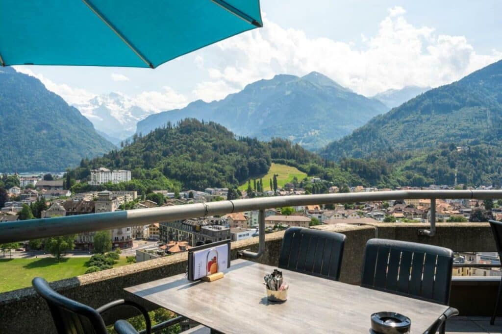 Imagem da varanda do Metropole Swiss Quality Hotel durante o dia com uma mesa quadrada a frente com três cadeiras, o local oferece vista para a cidade e montanhas.