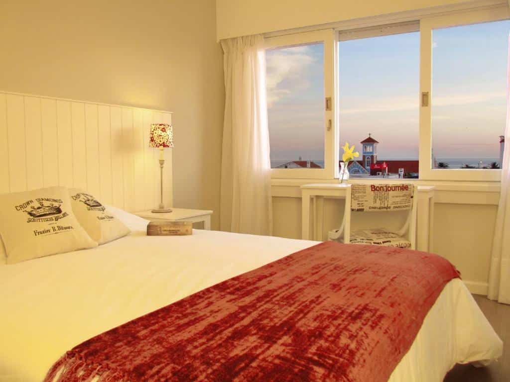 Uma cama com uma manta vermelha em um quarto bem iluminado no Atlántico Boutique Hotel (Península) com grandes janelas com vista para o oceano e um farol, para representar onde ficar em Évora.