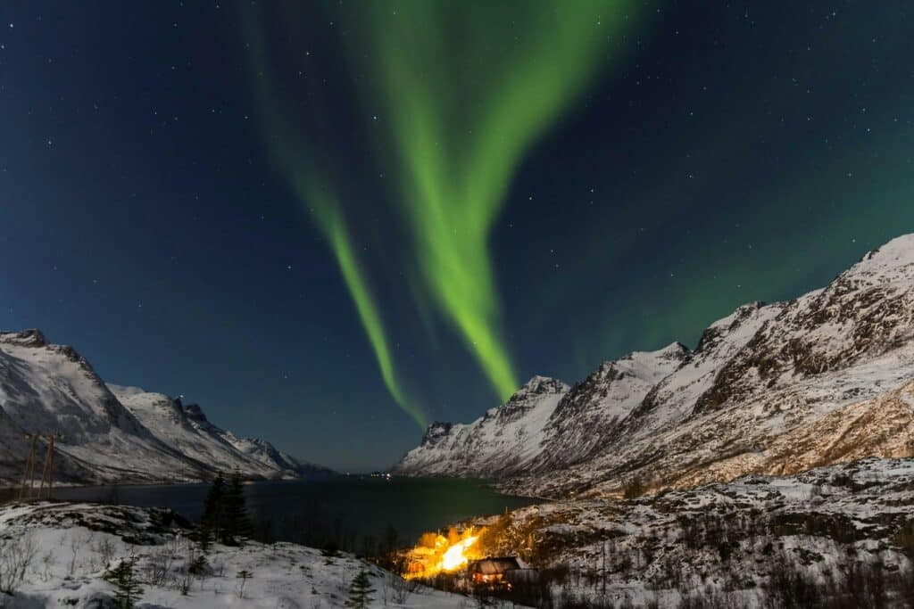 Aurora boreal ilumina o céu noturno acima de uma paisagem montanhosa coberta de neve em Tromø, na Noruega. Feixes de luz verde ondulante brilham intensamente contra o céu escuro estrelado, enquanto as montanhas e um lago tranquilo refletem o brilho suave.