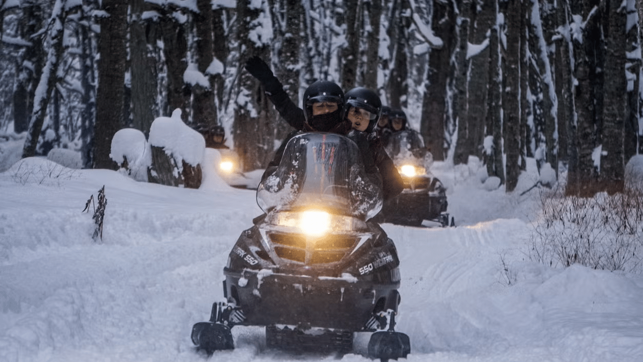 Duas pessoas em cima de uma moto na neve, no fundo árvores.