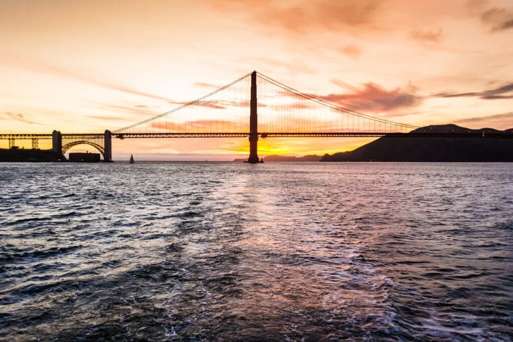 Foto da Ponte Golden Gate tirada durante o pôr do sol da perspectiva de um barco. É possível ver também o mar.