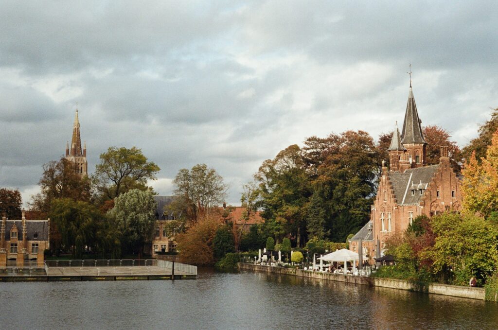 Paisagem de Bruges, na Bélgica, mostrando o canal cercado por edifícios históricos de tijolos vermelhos e torres pontiagudas. À esquerda, uma igreja gótica com uma torre alta e fina se destaca no cenário, enquanto à direita, há uma construção com torres menores, também medieval. As árvores outonais adicionam toques de laranja e verde ao cenário, e um pequeno cais com mesas e cadeiras oferece um local relaxante à beira da água.