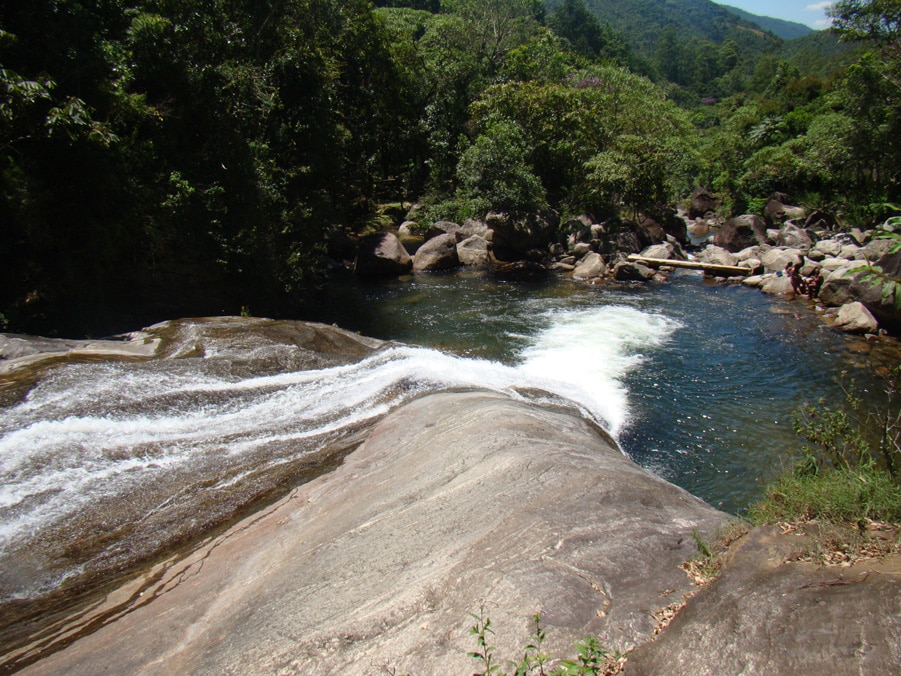Foto na parte de cima da Cachoeira do Escorrega que mostra a queda d'água formando um poço com várias pedras ao redor e algumas pessoas. Há também bastante árvores em volta durante o dia. Imagem ilustrando post O que fazer em Visconde de Mauá.