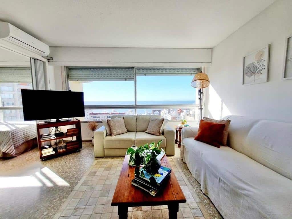 Vista para uma sala do Casatempo Apto Vista Mar em Punta Del Leste, Uruguai. Há dois sofás e uma mesa de centro. No lado esquerdo há uma estante com televisão. Atrás do sofá que está no centro, há uma janela com vista para o mar.