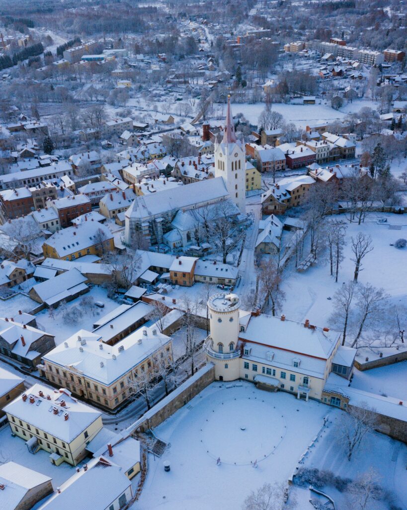 Vista aérea de Cesis, na Letônia, coberta de neve durante o inverno, com casas e ruas cercadas por árvores desfolhadas. No centro, destaca-se uma grande igreja com torre alta e pontiaguda.
