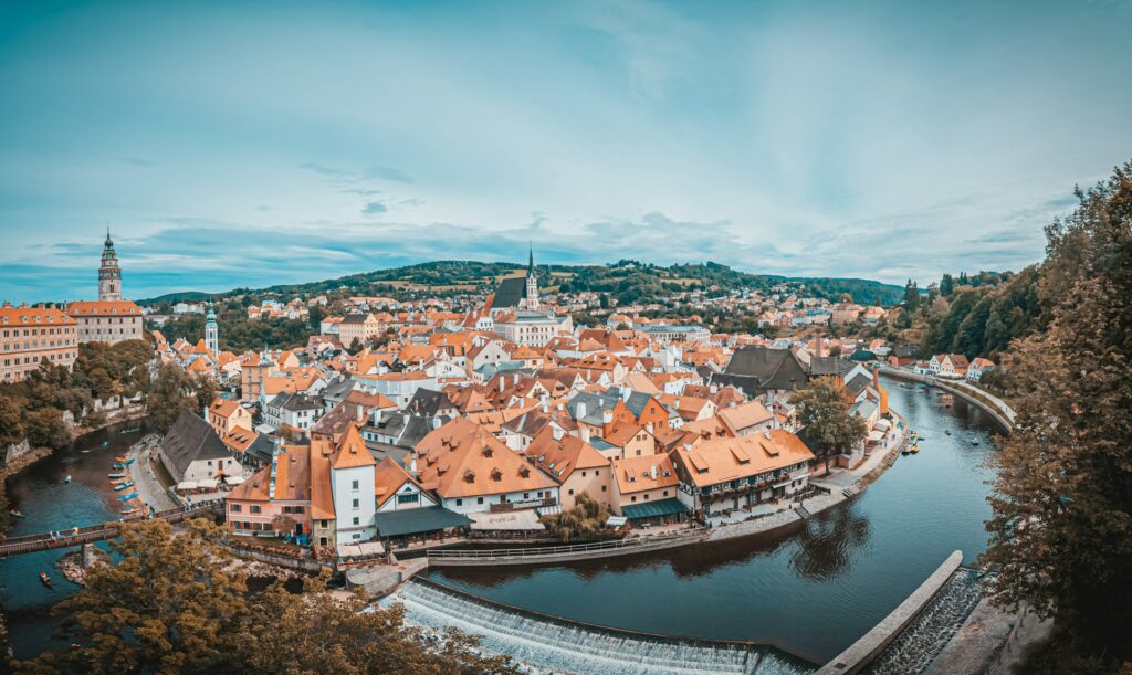 Panorâmica aérea de Český Krumlov, na República Tcheca, mostrando a cidade histórica com telhados laranja ao redor de um meandro do rio Vltava. Há vegetação verde e colinas ao fundo. No centro da imagem, destaca-se uma torre de igreja pontiaguda.