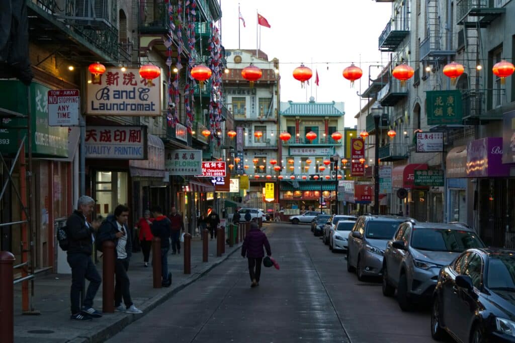 Foto de uma das ruas de Chinatown. Do lado esquerdo estão apenas lojas e pessoas passeando pela calçada. Do lado direito estão mais lojas e carros estacionados. Toda a rua é decorada com luzes vermelhas.