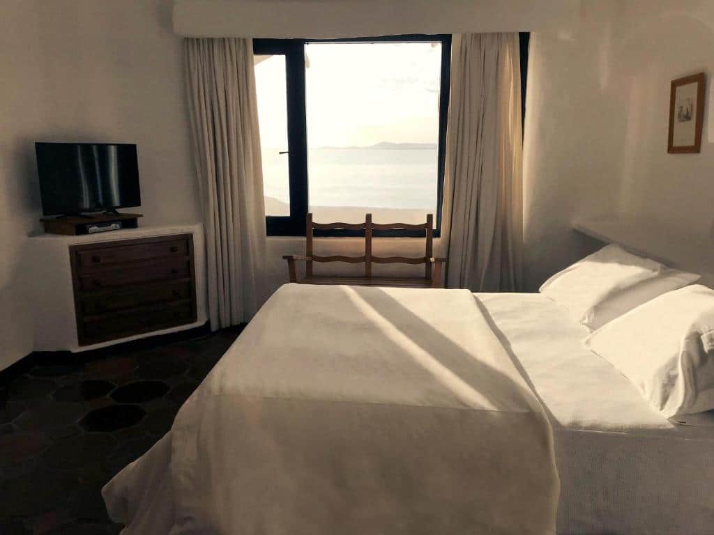 Uma cama bem arrumada no Club Hotel Casapueblo (Punta Ballena) em um quarto bem iluminado com janela, vista para o mar, TV, cômoda, banco de madeira e cortinas claras, para representar onde ficar em Punta Del Este.