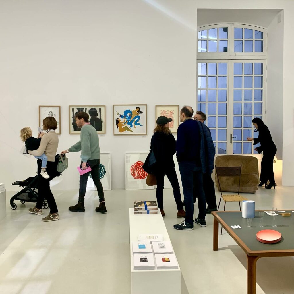 Imagem de pessoas andando na Collection Lambert Avignon com uma mesa do lado direito e uma banca no centro com panfletos e ao fundo pessoas olhando a exposição de quadro. Representa o que fazer em Avignon.