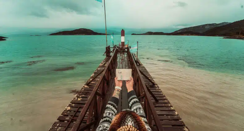píer no canal Beagle, em Ushuaia, com uma mulher ao centro segurando um passaporte aberto, mostrando a estampa do correio do fim do mundo. Ao final da passarela do píer há uma bandeira da argentina.