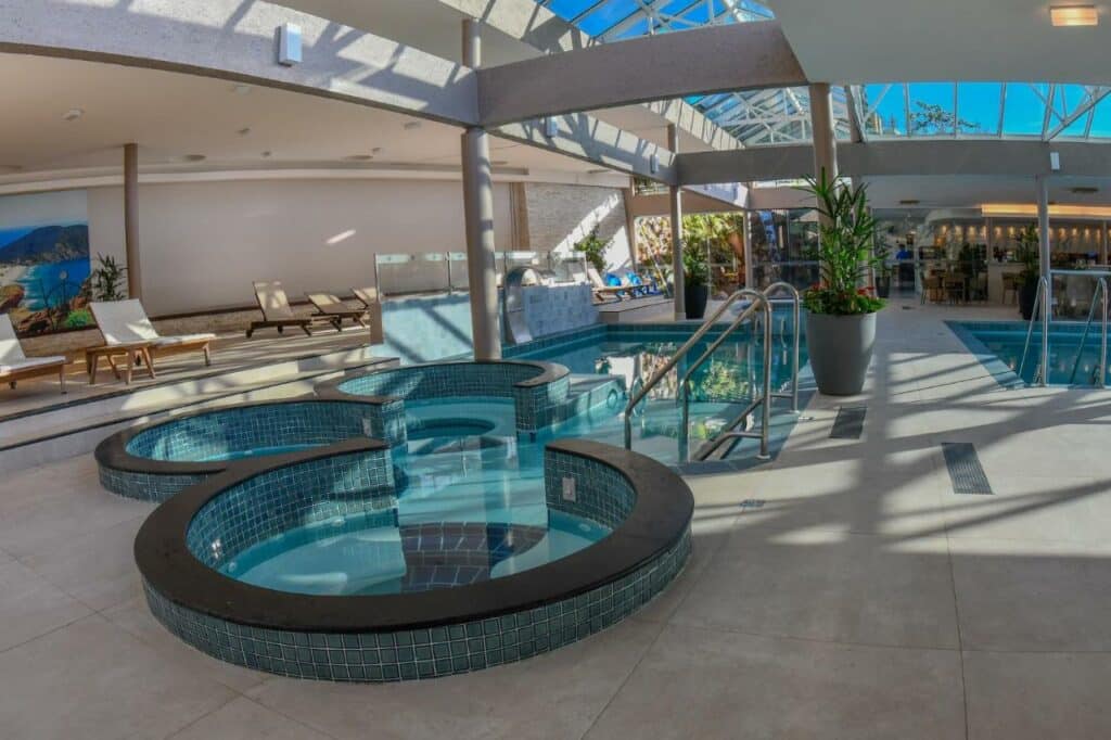 A imagem mostra uma piscina de hidromassagem em um ambiente luxuoso do Costão do Santinho Resort All Inclusive. Ao redor da piscina, há vegetação tropical exuberante e áreas de descanso com espreguiçadeiras confortáveis. O céu está claro e ensolarado, criando uma atmosfera relaxante e convidativa.