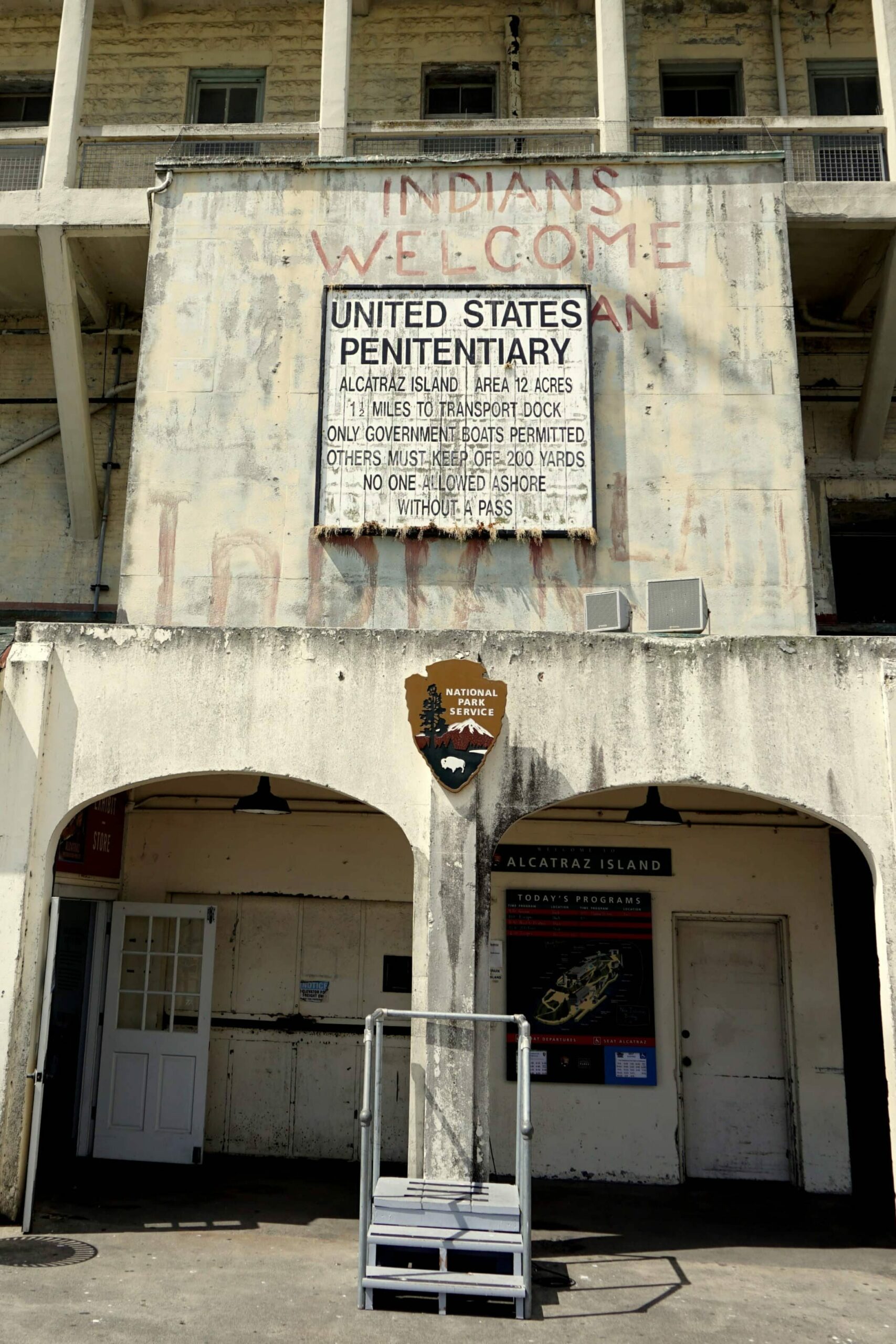 Entrada da prisão de Alcatraz, marcada por uma placa escrito "United States Penitentiary" e outras informações. A entrada é pequena, com duas divisões.