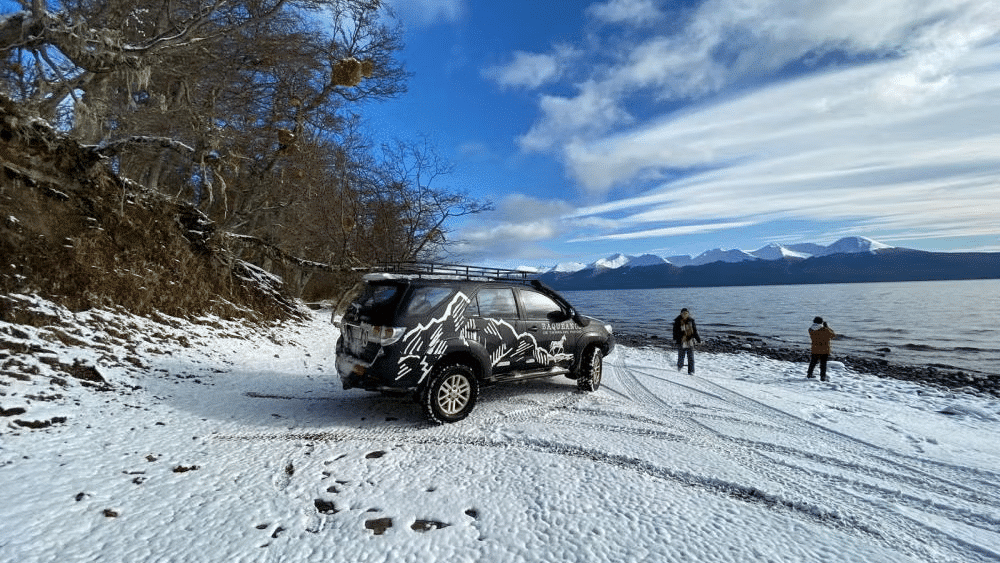 Um automóvel 4x4 parado na neve, do lado direito duas pessoas na beira do lago.Imagem para ilustrar post sobre passeios em Ushuaia.