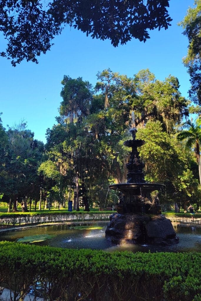 Uma fonte ornamentada jorra água em um pequeno lago cercado por uma cerca viva bem aparada. A fonte está situada em meio a um jardim exuberante, com árvores altas e densas que proporcionam sombra. O céu azul claro contrasta com o verde vibrante das folhas, criando uma atmosfera tranquila e serena.