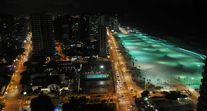 Vista aérea da cidade de Fortaleza. Está de noite então é possível ver as luzes da cidade. Enquanto os prédios estão iluminados a esquerda, à direita vemos a orla da Praia de Iracema iluminada.