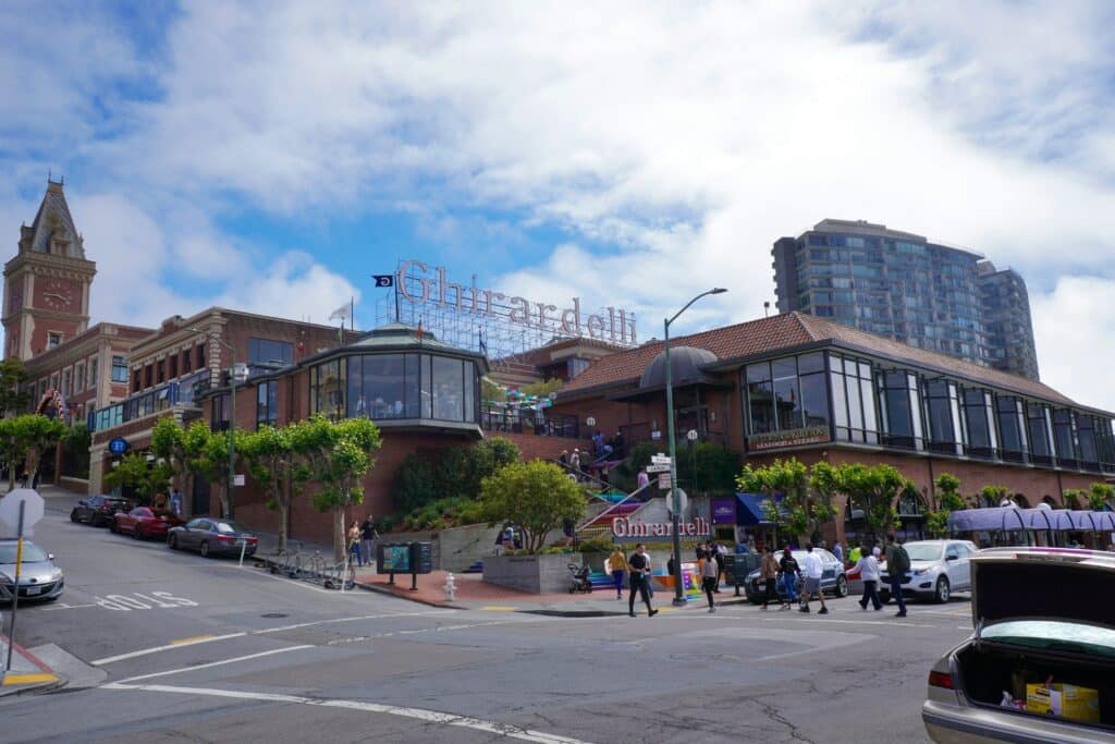 Vista da entrada da Ghirardelli. Uma placa bem grande acima do telhado indica o loca, e uma escada leva até a parte principal. Existem várias partes do prédio, que se estendem até outras partes da rua e da esquina.