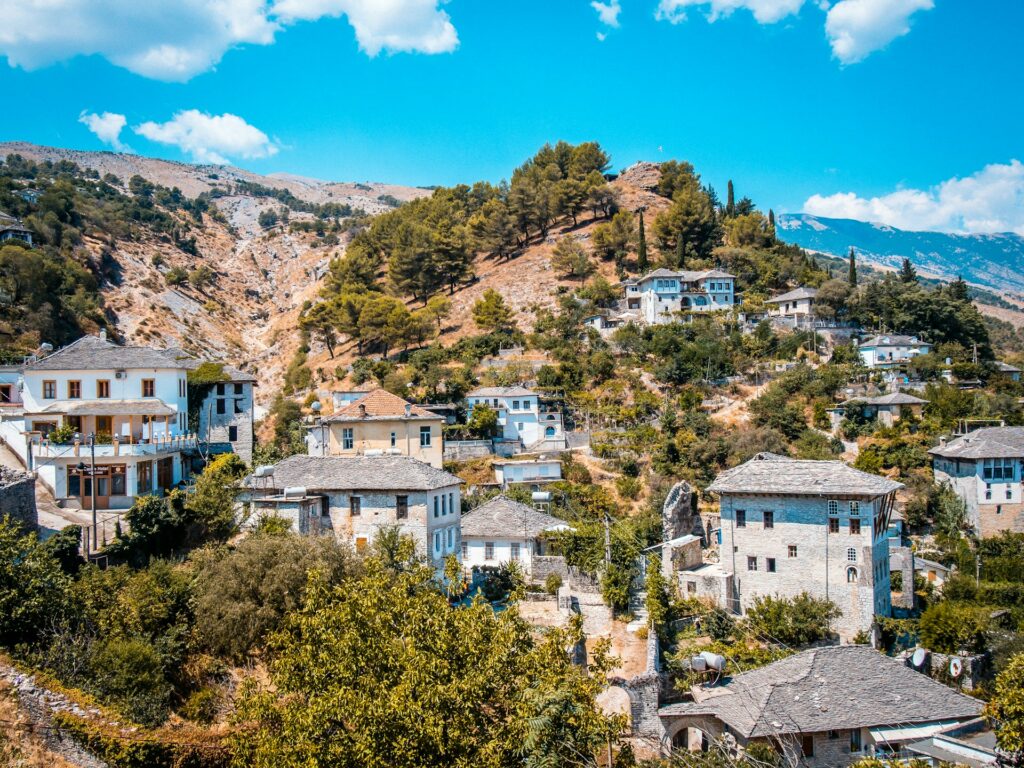 Uma aldeia de Gjirokastër, um dos destinos incomuns da Europa, encravada em encostas montanhosas, com casas de pedra com telhados cinzentos. A paisagem é pontuada por árvores e arbustos verdes, e as montanhas ao fundo são parcialmente cobertas por vegetação baixa. O céu é claro com algumas nuvens esparsas.