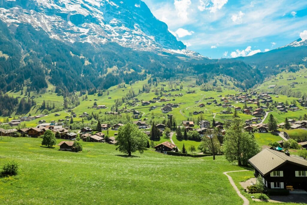 Imagem durante o dia da cidade de Grindelwald com casinhas espelhadas pelo local em meio a árvores e ao fundo da cidade montanhas. Representa o que fazer na Suíça.