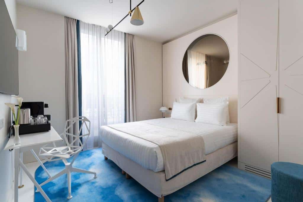 A imagem mostra um quarto moderno e bem iluminado no Hotel Astra Opera - Astotel, um dos hotéis 4 estrelas em Paris. A cama é grande, com roupa de cama branca e dois travesseiros, posicionada contra uma parede clara com um grande espelho redondo acima da cabeceira. O quarto possui uma janela alta com cortinas translúcidas, permitindo a entrada de luz natural. Há um tapete azul claro no chão, uma cadeira branca de design geométrico e uma mesa com um conjunto de café. A decoração é minimalista e elegante, criando um ambiente tranquilo e acolhedor.