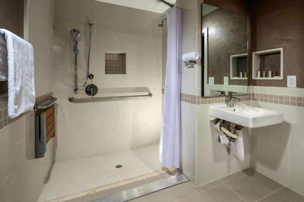 Banheiro do Hotel Aventura. Do lado direito uma pia e um espelho. Do lado esquerdo um box com cadeira, barras de apoio, chuveiro e mangueira.