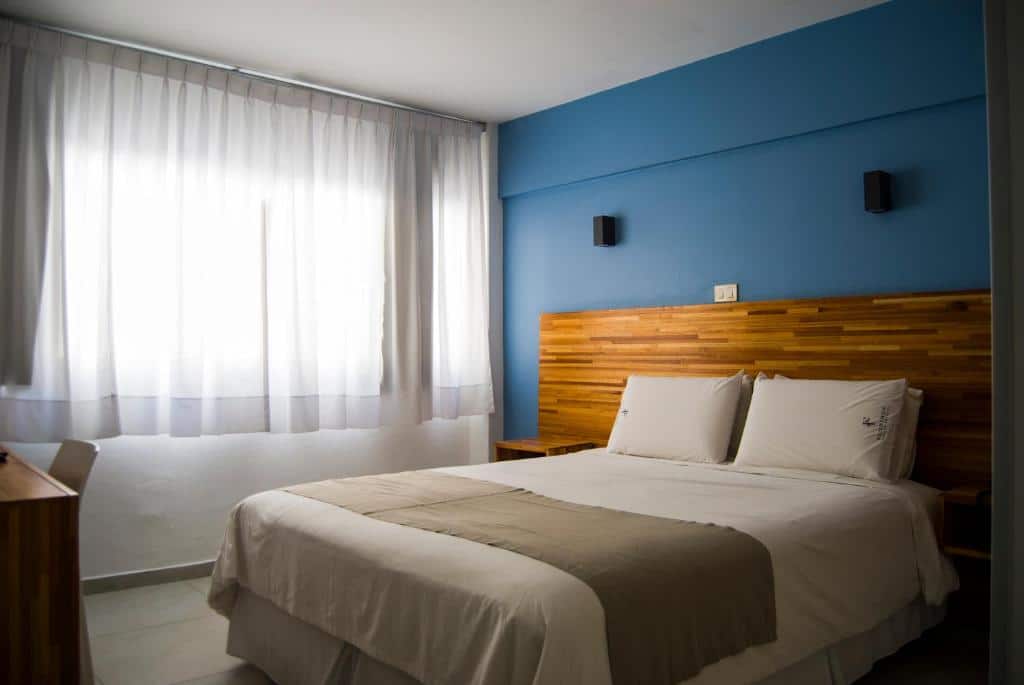 Uma cama de casal bem arrumada em um quarto simples no Hotel Florinda, com paredes azuis, detalhes em madeira e luz do sol entrando por cortinas transparentes, para representar onde ficar em Punta Del Este.