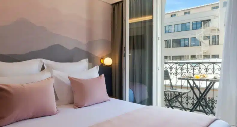 Quarto do Hôtel Le Milie Rose, um dos hotéis para brasileiros em Paris. Uma cama de casal está ao lado esquerdo da imagem com mesinhas de cabeceira e luminárias dos dois lados. Ao lado direito há uma porta aberta para a varanda que tem vista para prédios na vizihança.