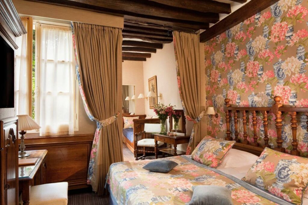 Quarto do Hotel Left Bank Saint Germain, um dos hotéis 3 estrelas em Paris. Uma cama de casal está ao lado direito da imagem com luminárias dos dois lados. Em frente fica uma mesa de trabalho com cadeira, abajur e TV na parede acima. Na lateral esquerda da cama fica uma área de estar com poltrona e sofá. Os cômodos são separados por uma cortina.