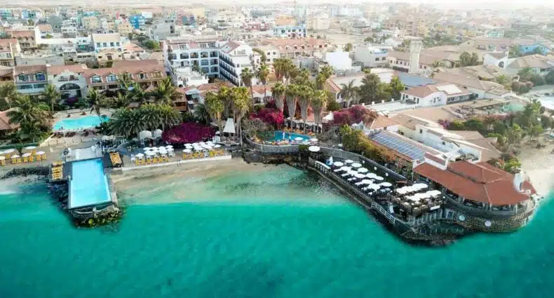 Vista aérea do Hotel Odjo d'água com piscinas, palmeiras e edifícios ao lado de água azul-turquesa clara, para representar hotéis em Cabo Verde.