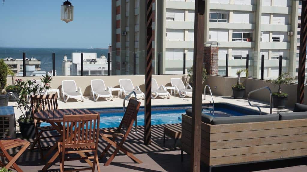 Área externa na cobertura com uma pequena piscina, espreguiçadeiras, móveis de madeira e prédios da cidade ao fundo, na propriedade do Remanso.
