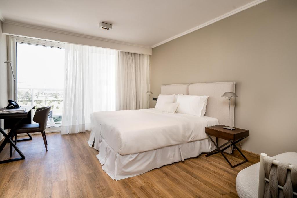 Quarto do Hotel Solerios, o ambiente é espaçoso e há uma cama box de casal no centro, com lençóis brancos e uma cabeceira bege atrás. Está ladeada por dois criados-mudos. Ao lado da cama há uma grande janela com cortinas brancas, parra representar onde ficar em Punta Del Este.