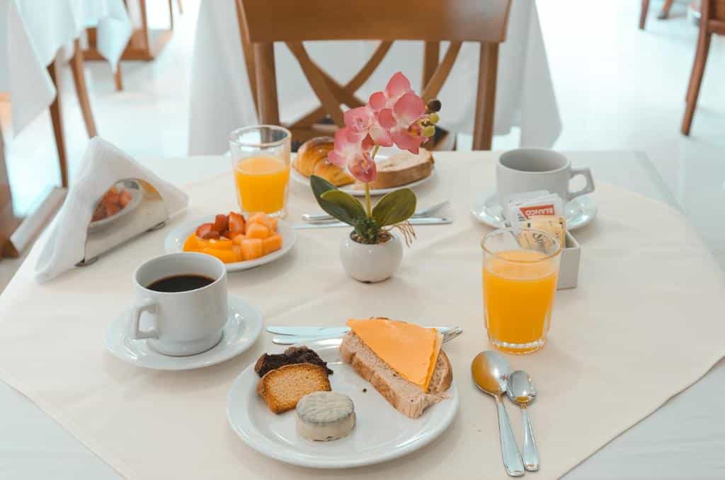 Uma mesa de café da manhã com café, suco de laranja, pão, queijo, frutas, doces e uma pequena orquídea em vaso, servido no Hotel Sunset Beach (Praia Mansa).